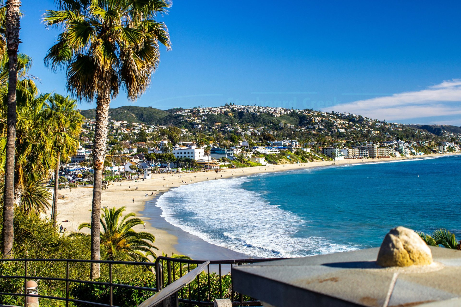 Scenic coastal beach view with palm trees, sandy shore, turquoise waves, hillside homes, and clear blue sky